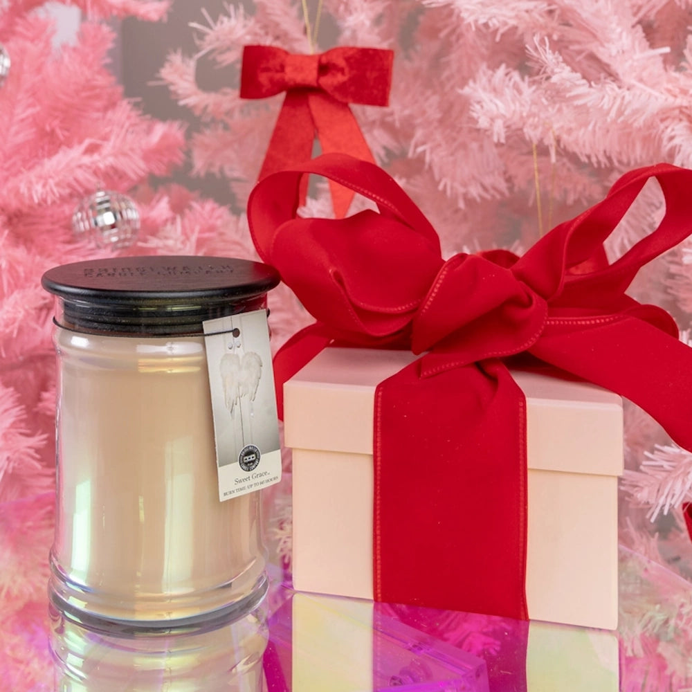 A glass jar candle next to a gift box with a red bow, set against a pink Christmas tree background.