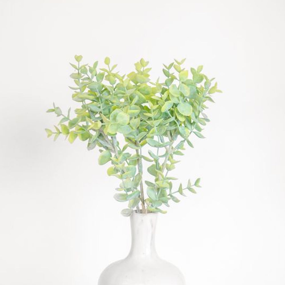 Green eucalyptus stems with round leaves in a white marble-patterned vase against a plain white background.