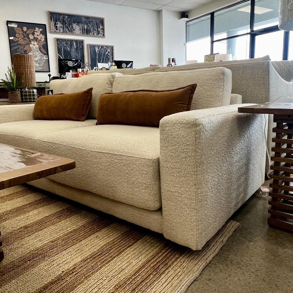 Cream textured sofa with brown lumbar pillows displayed in a showroom with a woven rug and coffee table.