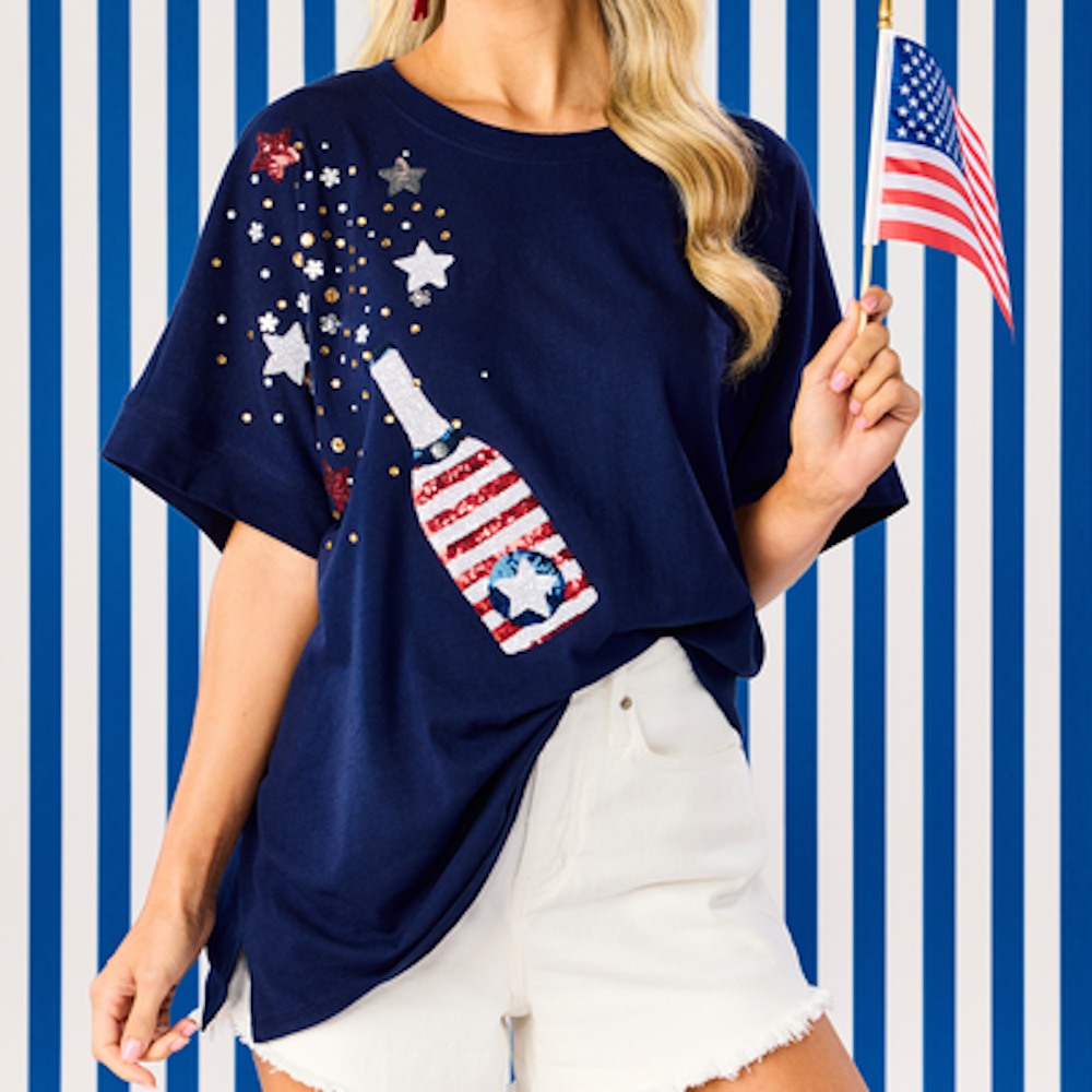 Woman in a navy shirt with a sequined champagne bottle and stars design, holding a small American flag.