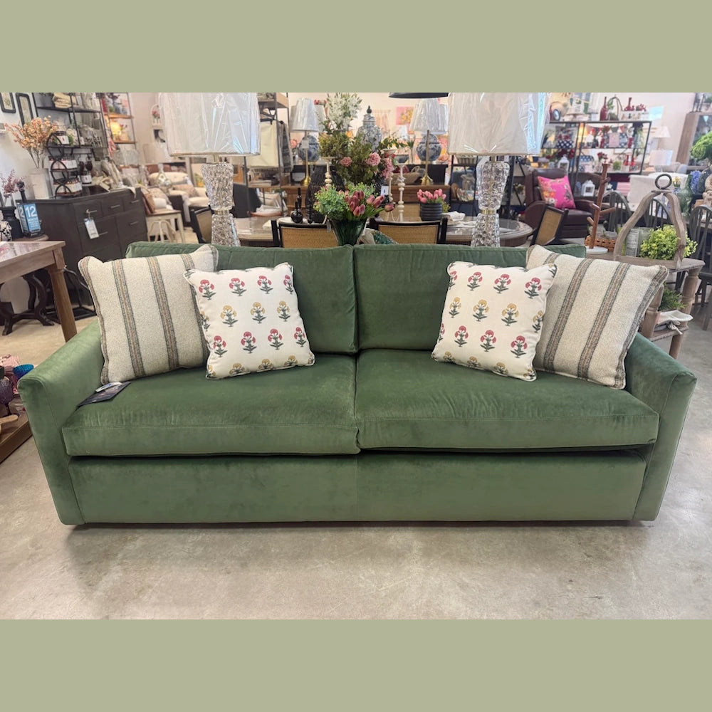 Emerald green velvet sofa with striped and floral throw pillows, displayed in a well-lit home furniture showroom setting.