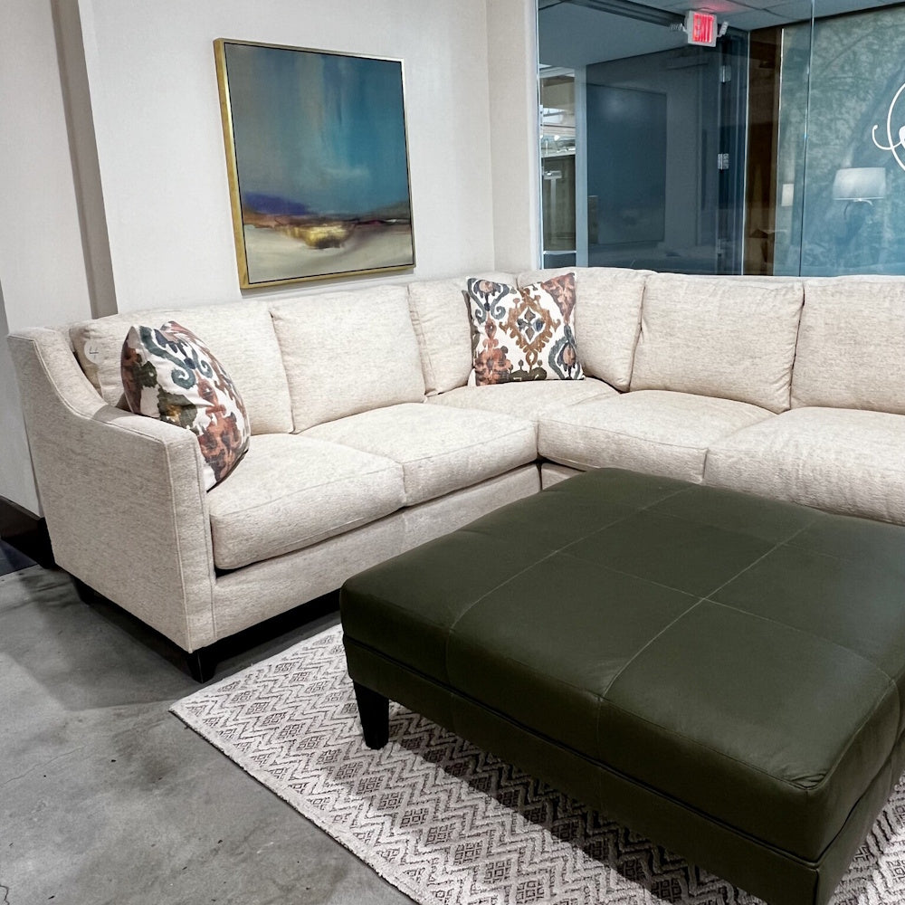 Beige L-shaped sectional couch with patterned pillows and a dark green leather ottoman on a rug.