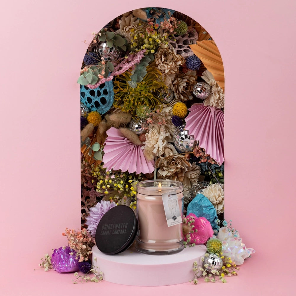 Lit glass jar candle displayed on a pedestal in front of an arch filled with dried flowers and disco balls.