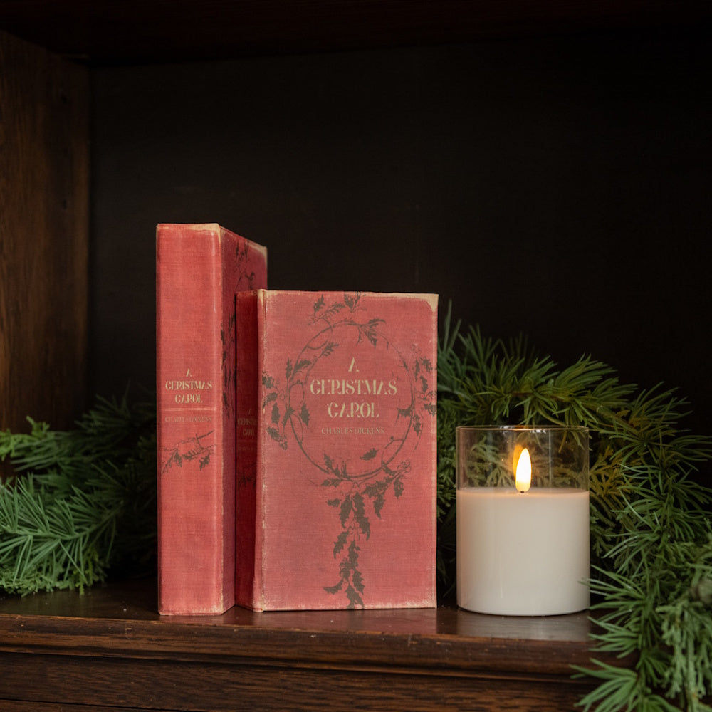 Two vintage red books titled "A Christmas Carol" beside a burning candle and evergreen pine garland on a shelf.