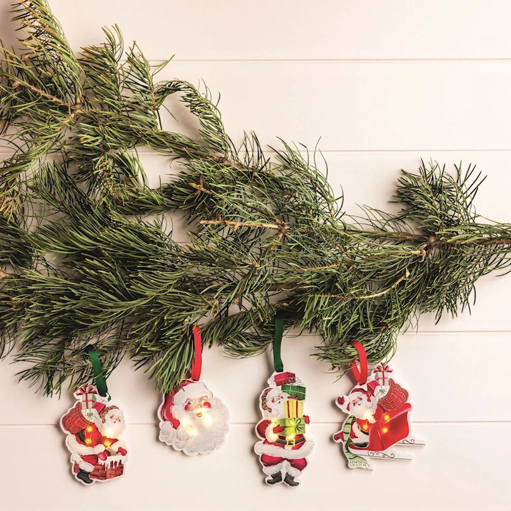 Four LED Santa Claus ornaments hanging from a pine branch against a white wooden background.