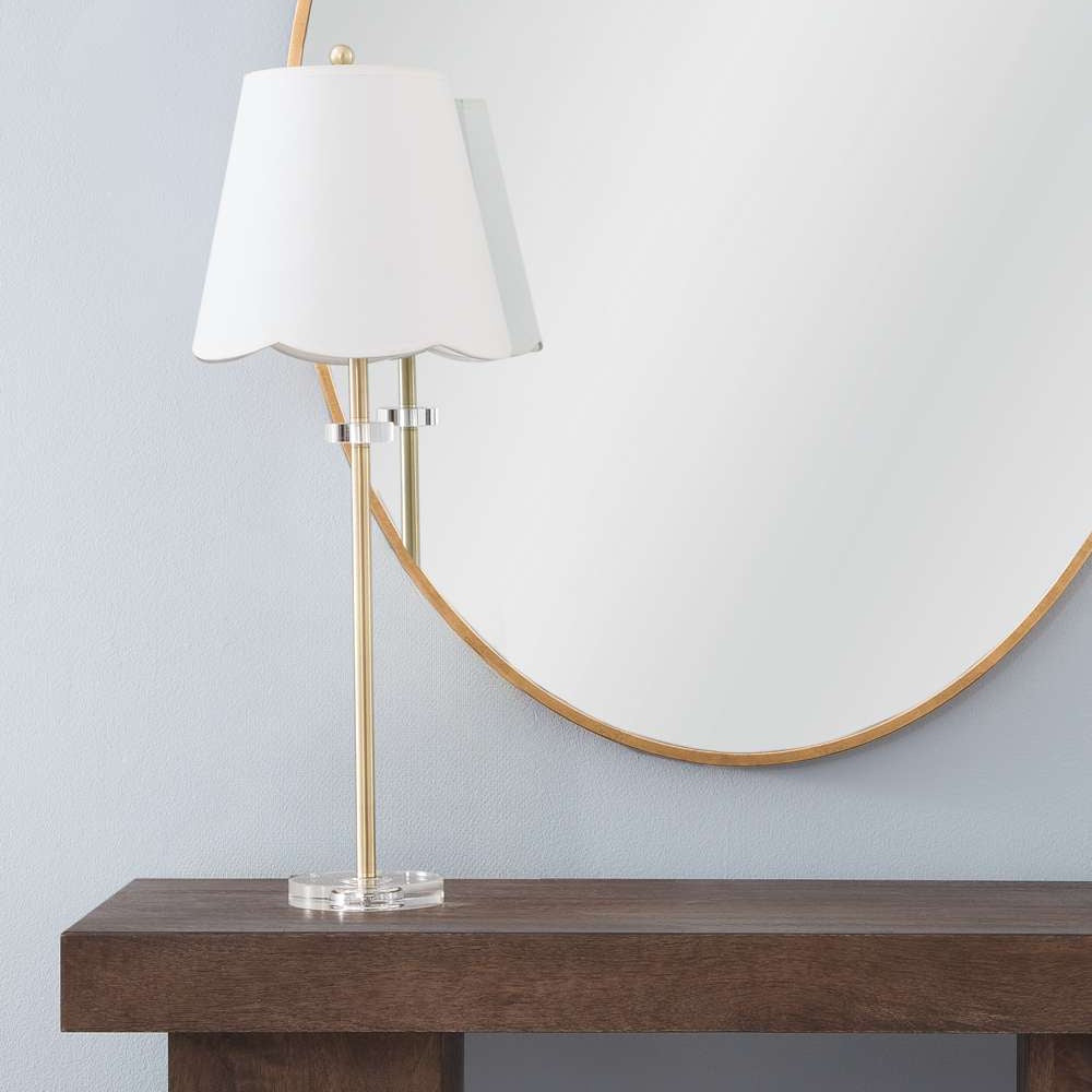 Modern brass table lamp with scalloped white shade on a wooden console table in front of a round gold mirror.