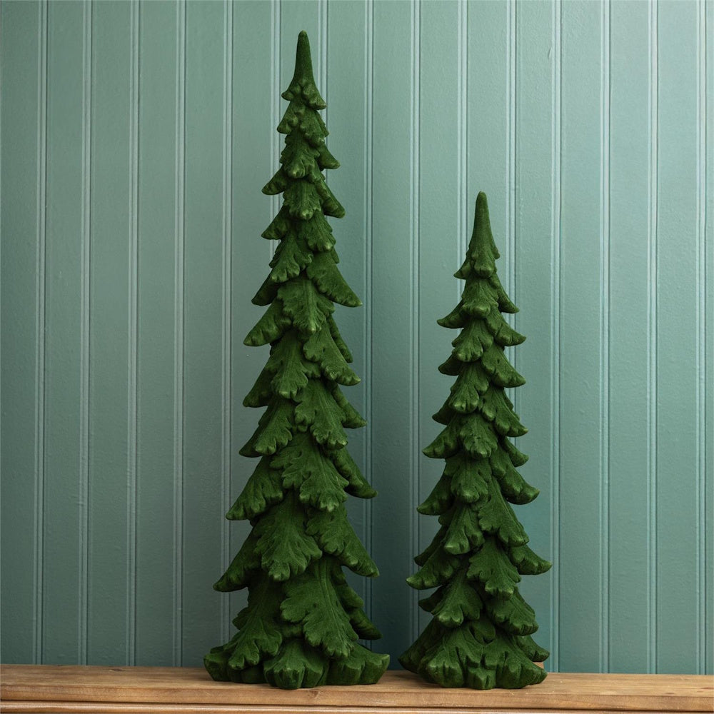 Two tall, slender green velvet decorative pine trees sitting on a wooden surface against a wall.