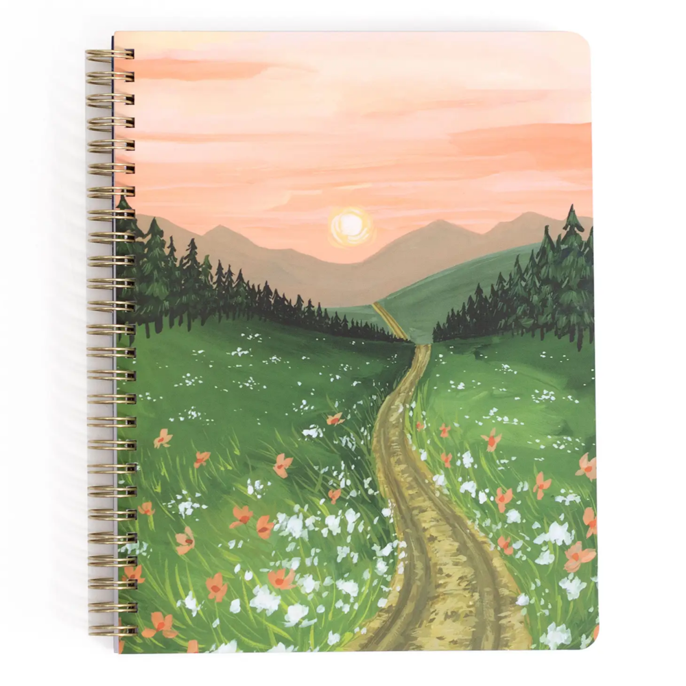 A spiral notebook featuring a painted landscape of a dirt path through a flowery meadow at sunset.