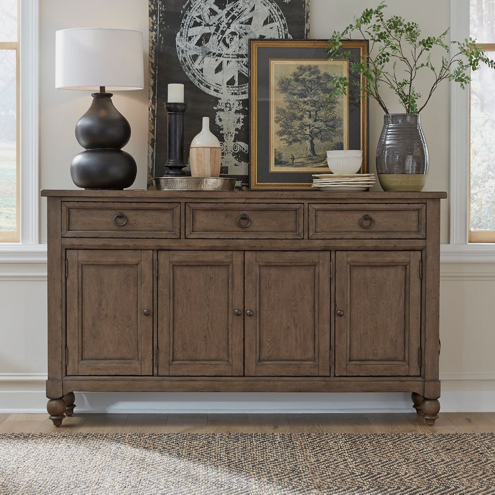Brown wooden farmhouse buffet styled with a table lamp, framed artwork, and green branches.