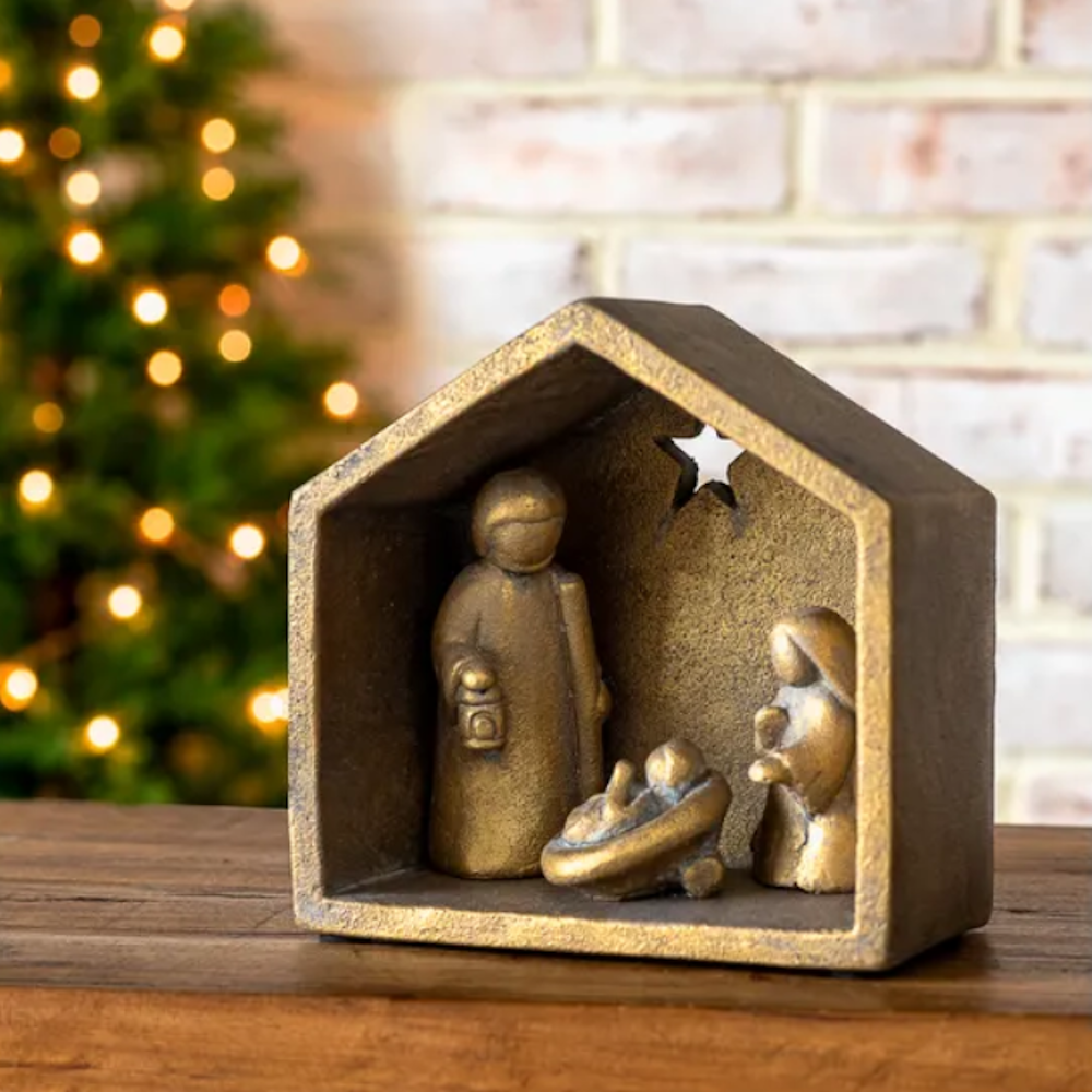 Antique gold Nativity scene in a house-shaped frame with a lit Christmas tree in the background.