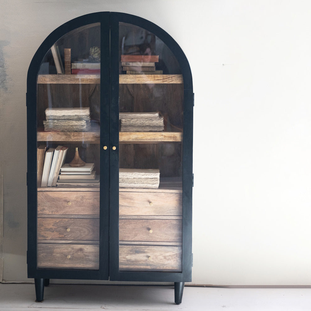 Arched black mango wood cabinet with two glass doors, wood shelves, and drawers, filled with books and stationery.