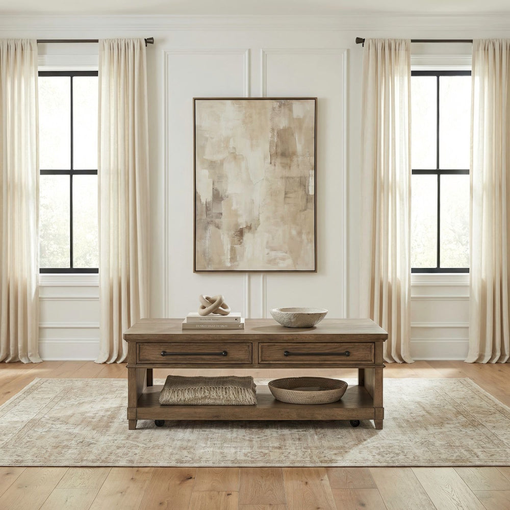 Rectangular wooden coffee table styled on a patterned beige rug with draped windows and abstract art.