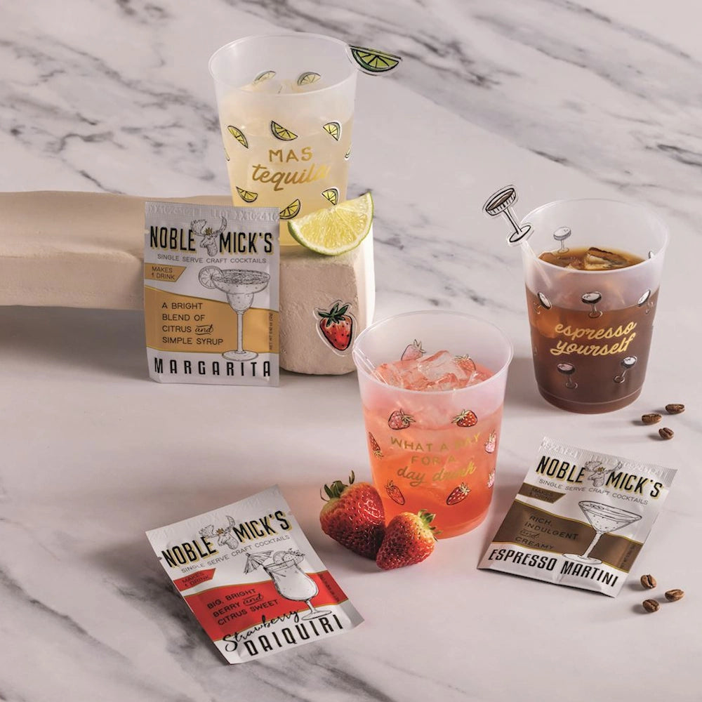 Three frosted cocktail cups with drinks alongside three Noble Mick's single-serve craft cocktail packets.