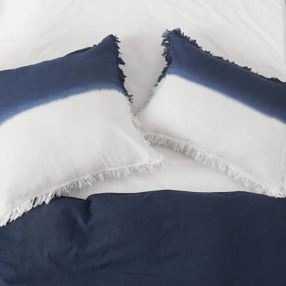 Two blue and white dip-dye ombre pillows with fringed edges on a bed.