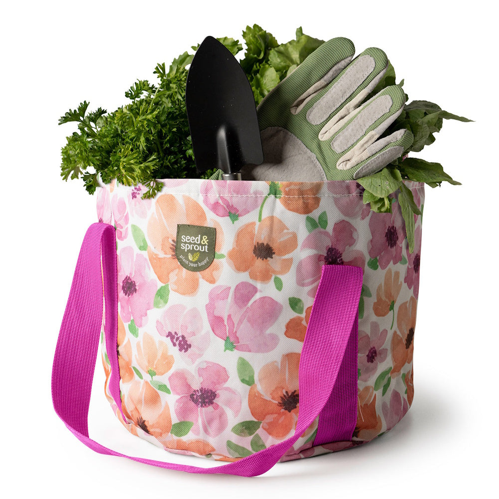 A floral canvas gardening tote filled with leafy green plants, a black hand trowel, and garden gloves.