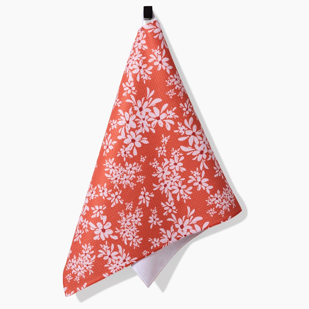 A hanging orange tea towel with a white floral pattern and a textured white underside against a plain backdrop.