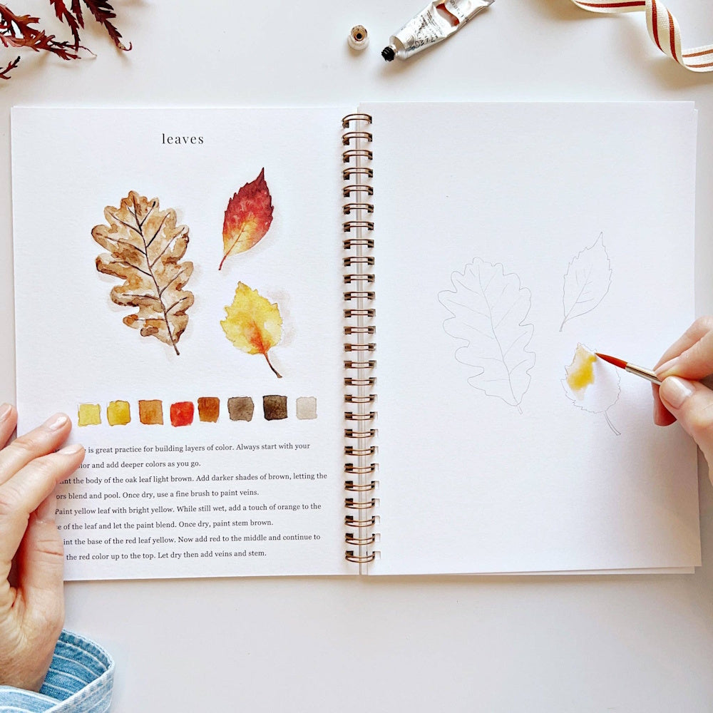 Open autumn watercolor workbook showing fall leaf illustrations and a hand painting a yellow leaf sketch.