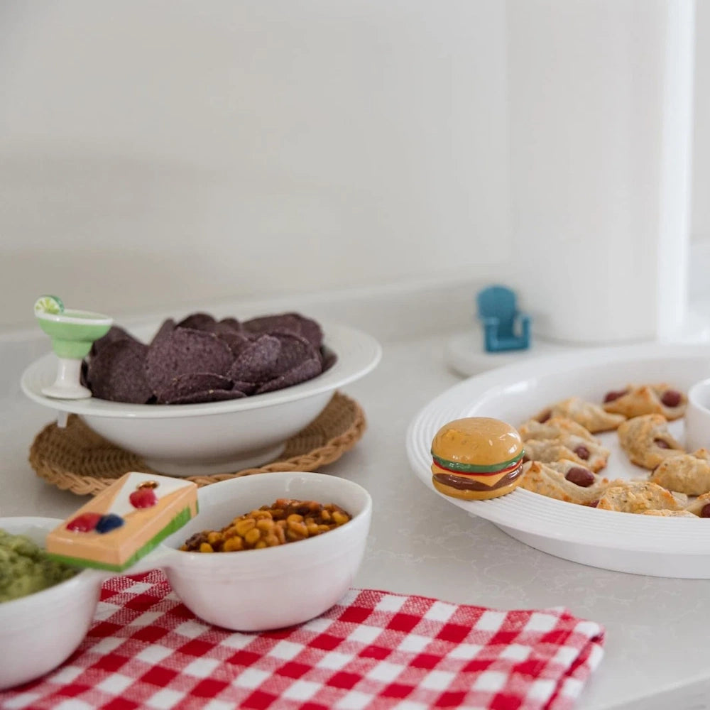 Appetizer spread with blue corn chips, pigs in a blanket, and corn dip, featuring decorative food-themed dish clips.