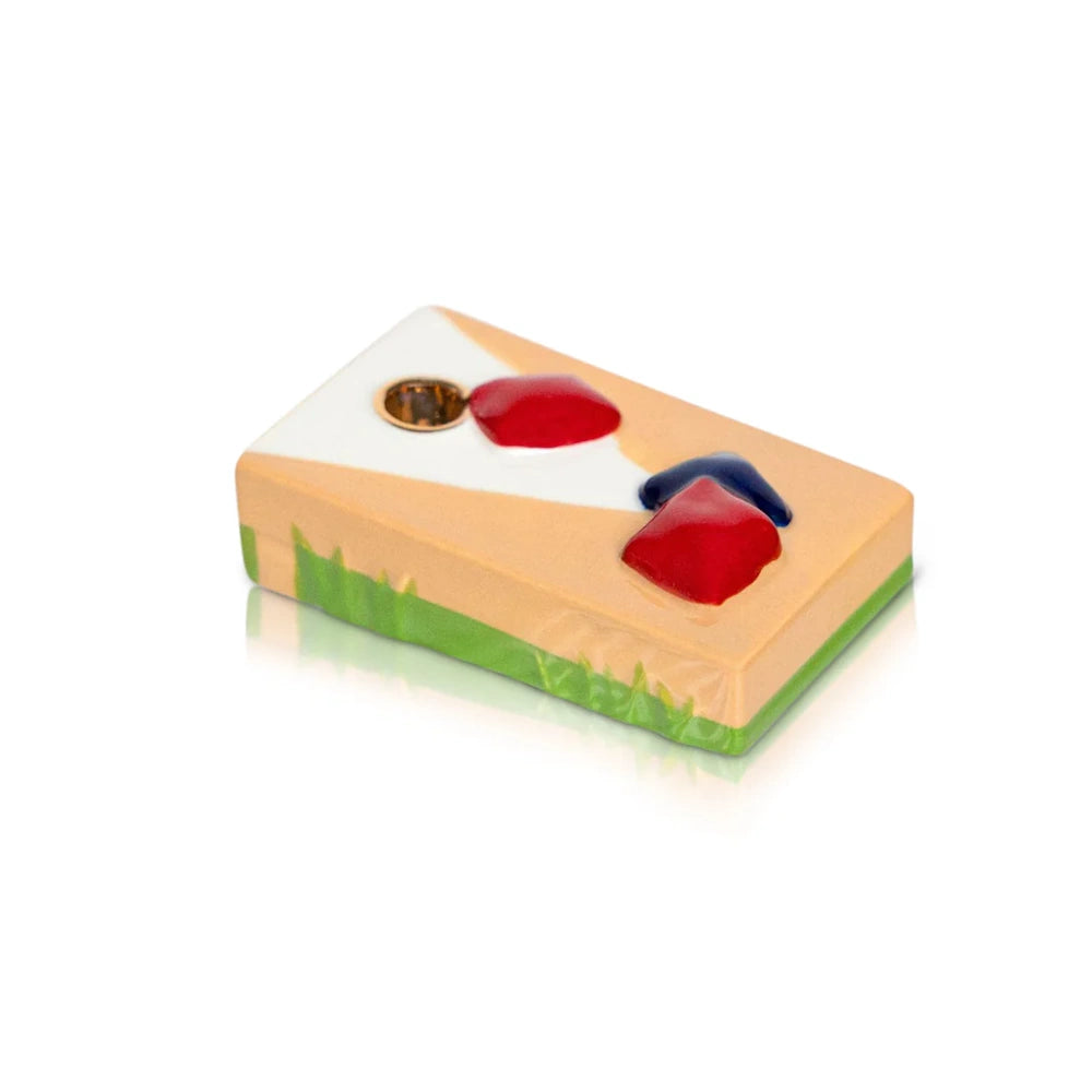Colorful cornhole board enamel pin featuring red and blue bean bags on a wooden board with green grass accents.