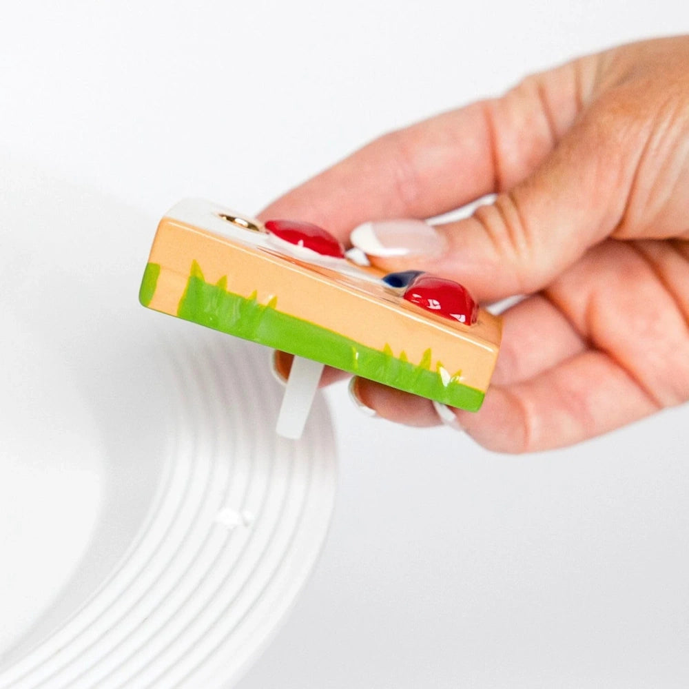 A hand inserts a colorful "Baggin' Rights" cornhole board ceramic garnish into the rim of a white serving platter.