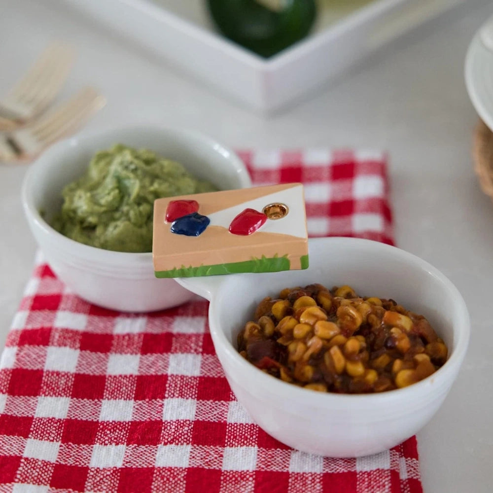 Miniature cornhole board decorative garnish on a white dip bowl with guacamole and salsa on a red checkered napkin.