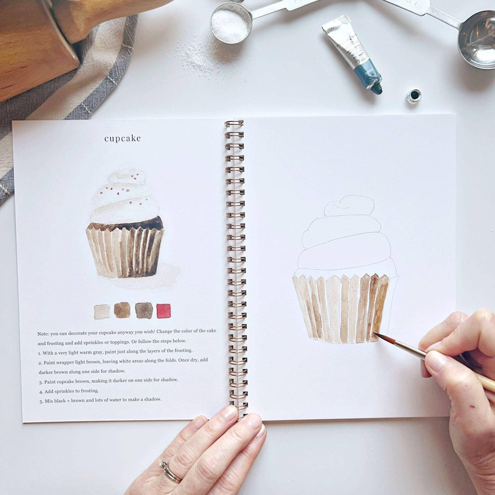 Watercolor workbook open to a cupcake tutorial with hands painting a brown cupcake liner using a fine brush.