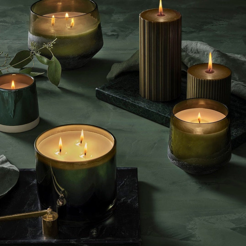 A collection of lit candles in various green glass jars and textured pillars on a dark surface.