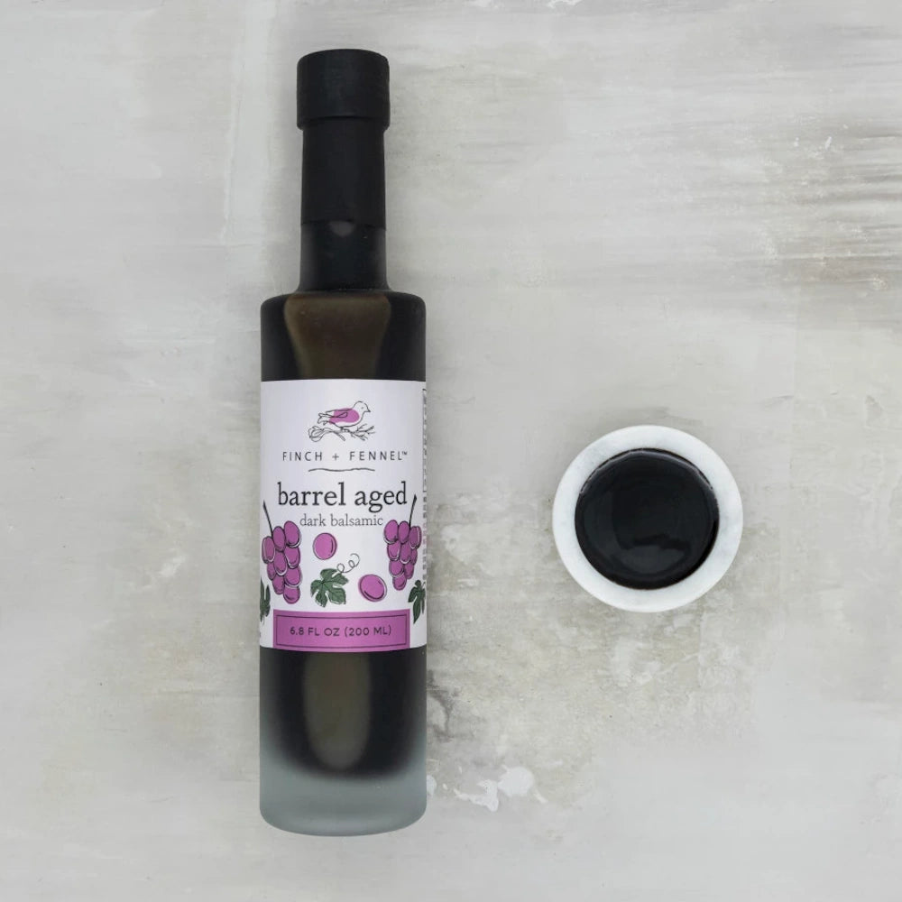 Finch + Fennel barrel aged dark balsamic vinegar bottle and a small dipping bowl of vinegar on a gray stone surface.