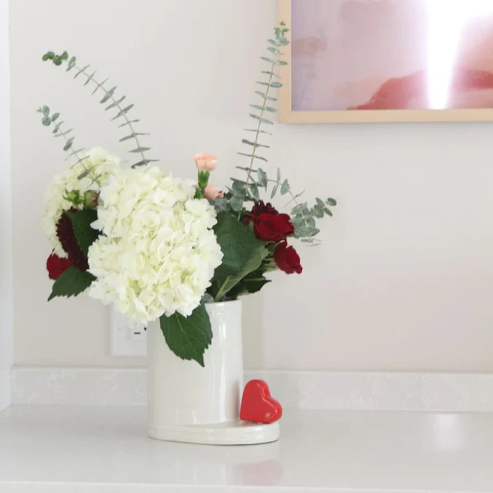 A floral bouquet with white hydrangeas and red roses in a white vase next to a small red heart.