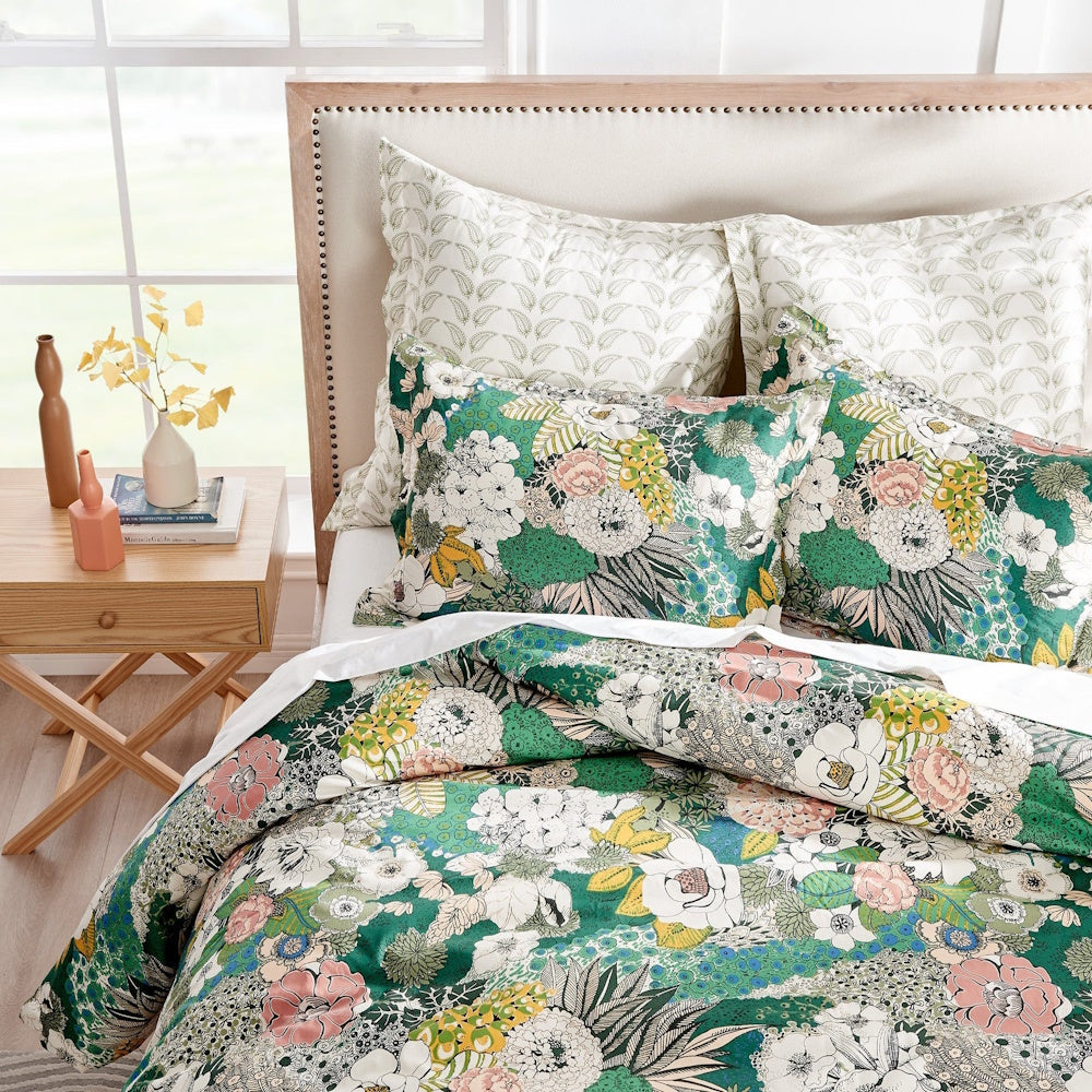 A bed styled with a green botanical comforter and pillow shams features a modern headboard and a wooden nightstand.