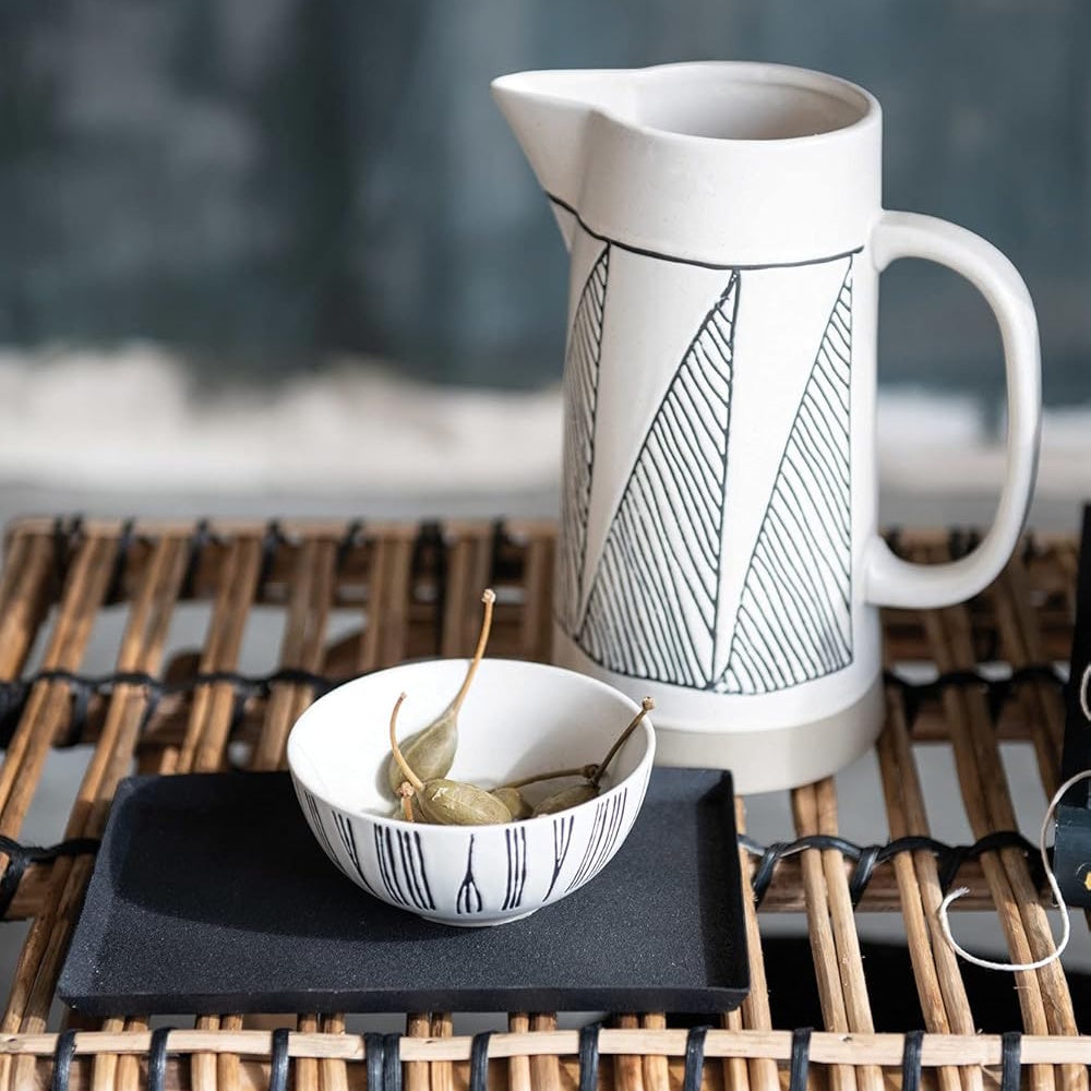 A cream stoneware pitcher with black geometric patterns next to a small bowl on a wicker surface.