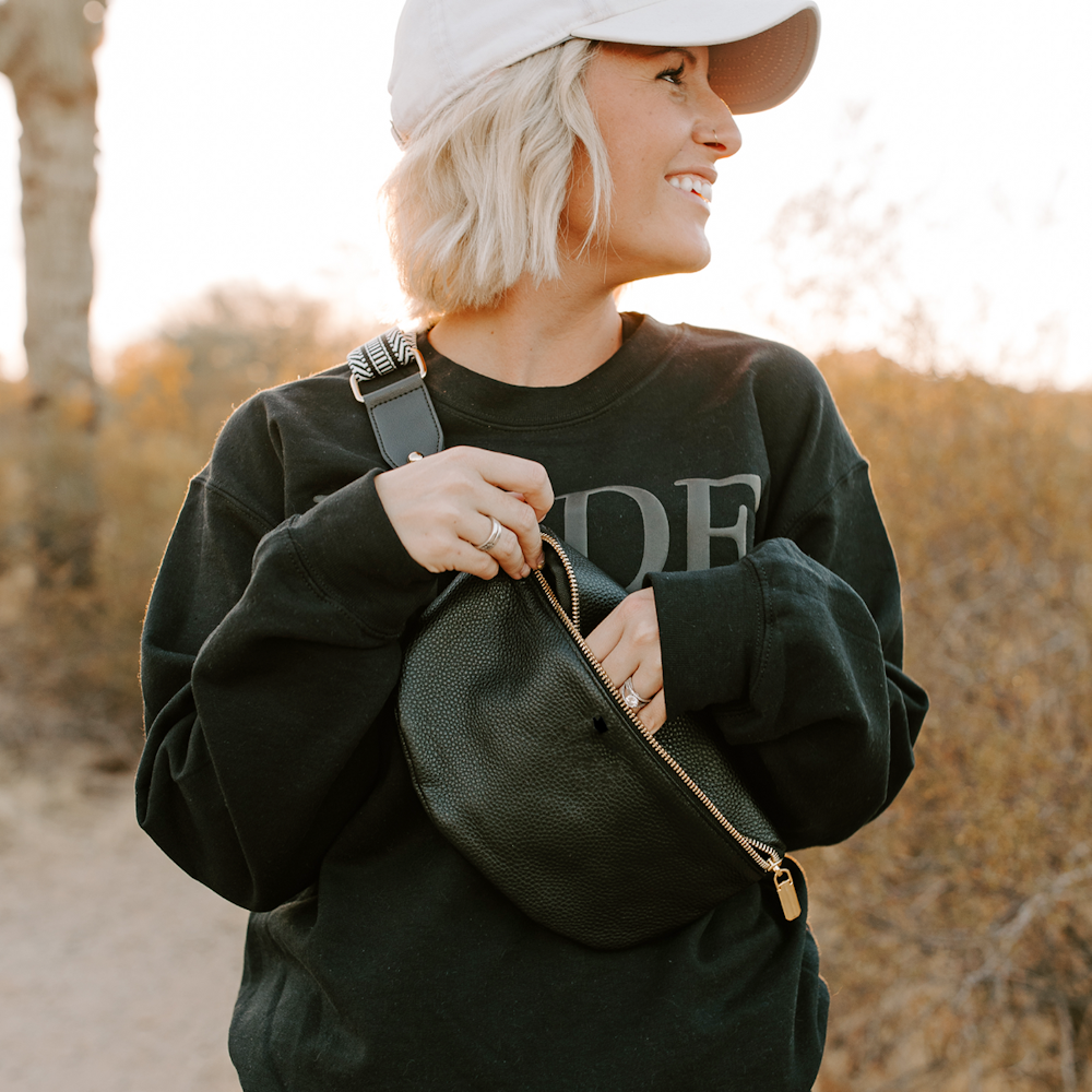 Woman with blonde hair smiling while reaching into a black leather crossbody fanny pack with gold hardware.