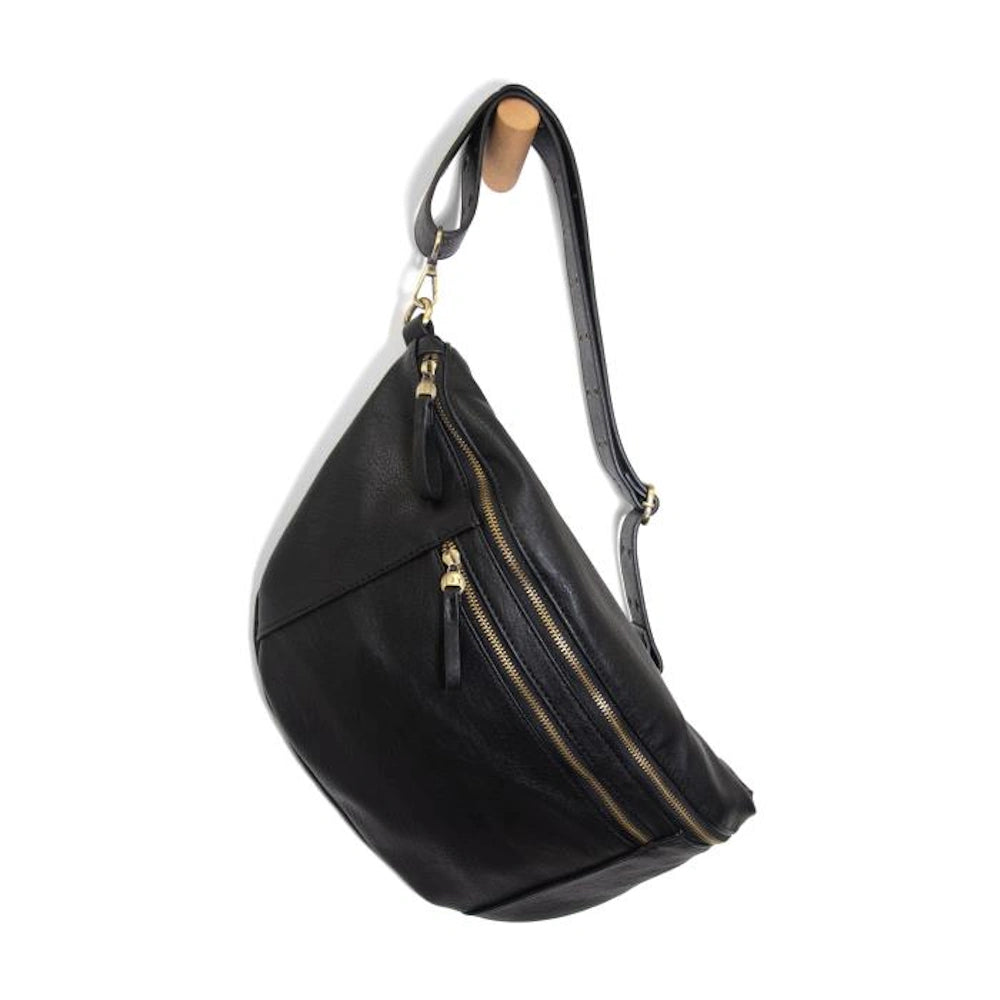 Black leather crossbody sling bag with gold zippers and adjustable strap hanging on a wooden wall peg.