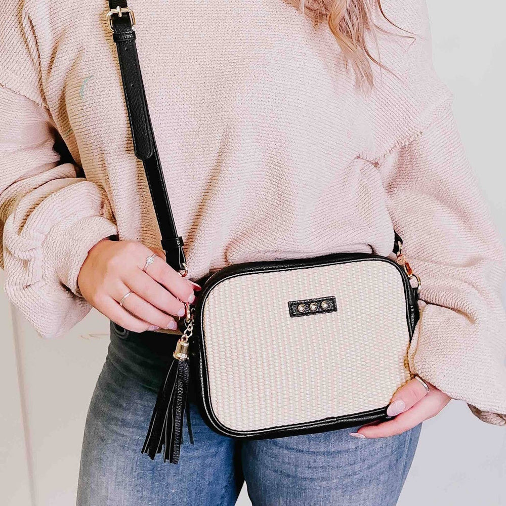 A person wearing a beige sweater and jeans holds a cream and black rectangular crossbody bag with a tassel.