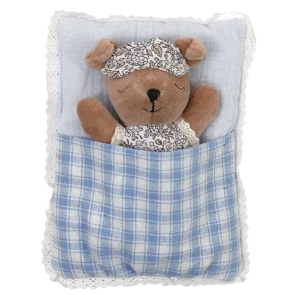 Small plush bear wearing a patterned eye mask sleeping in a blue plaid sleeping bag with lace trim.