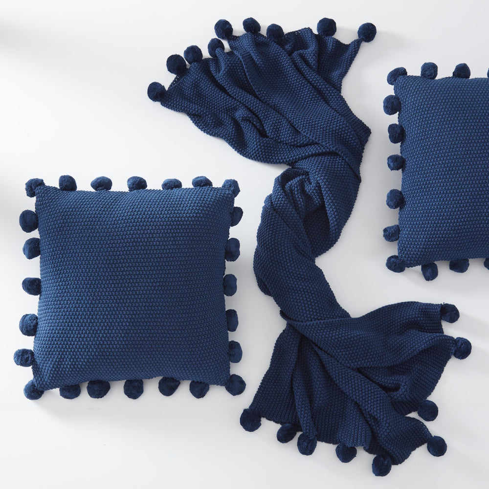 Navy blue moss stitch knit throw blanket and matching pillows with pompom trim on a white background.