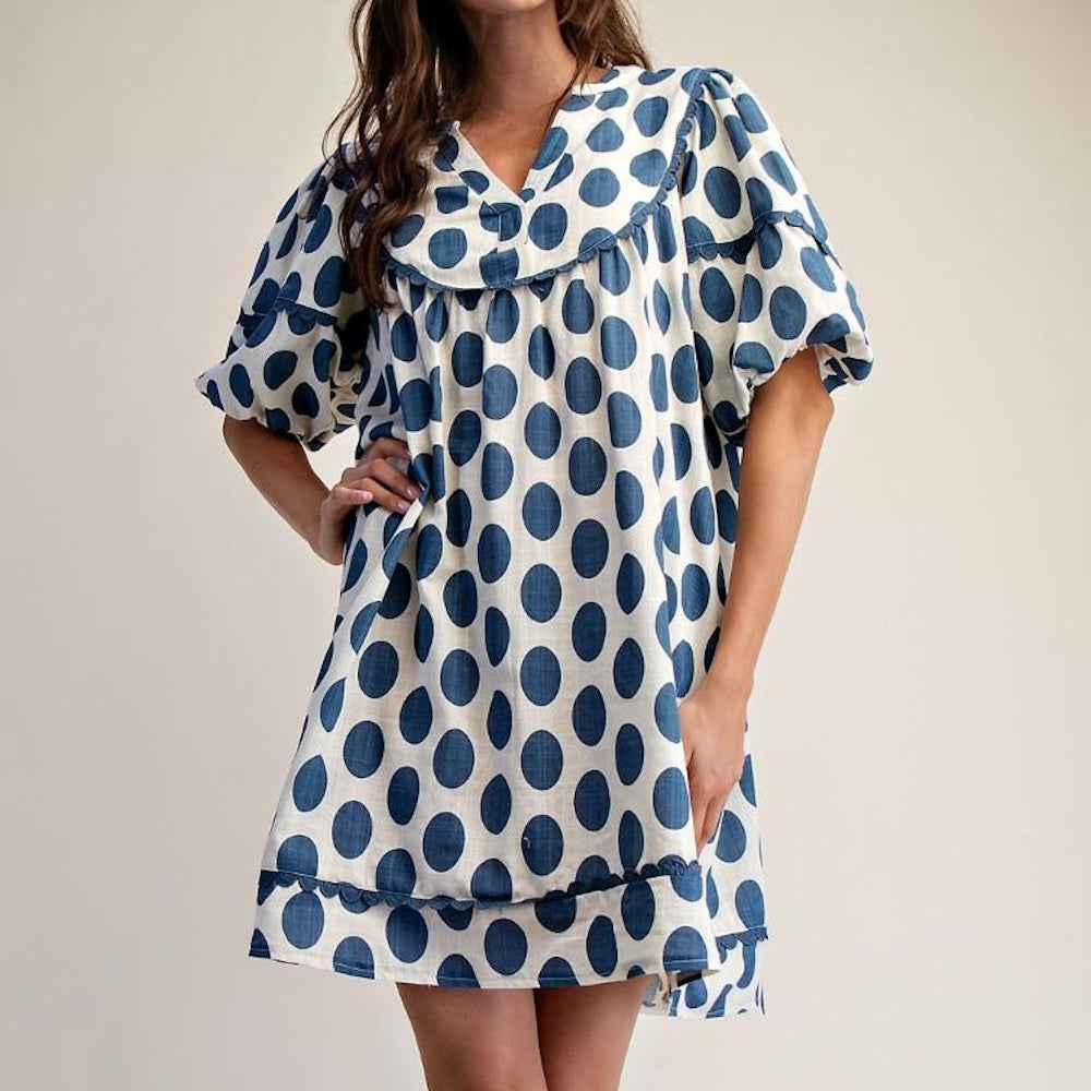 Woman wearing a white mini dress with large blue polka dots, a V-neckline, and puff sleeves.