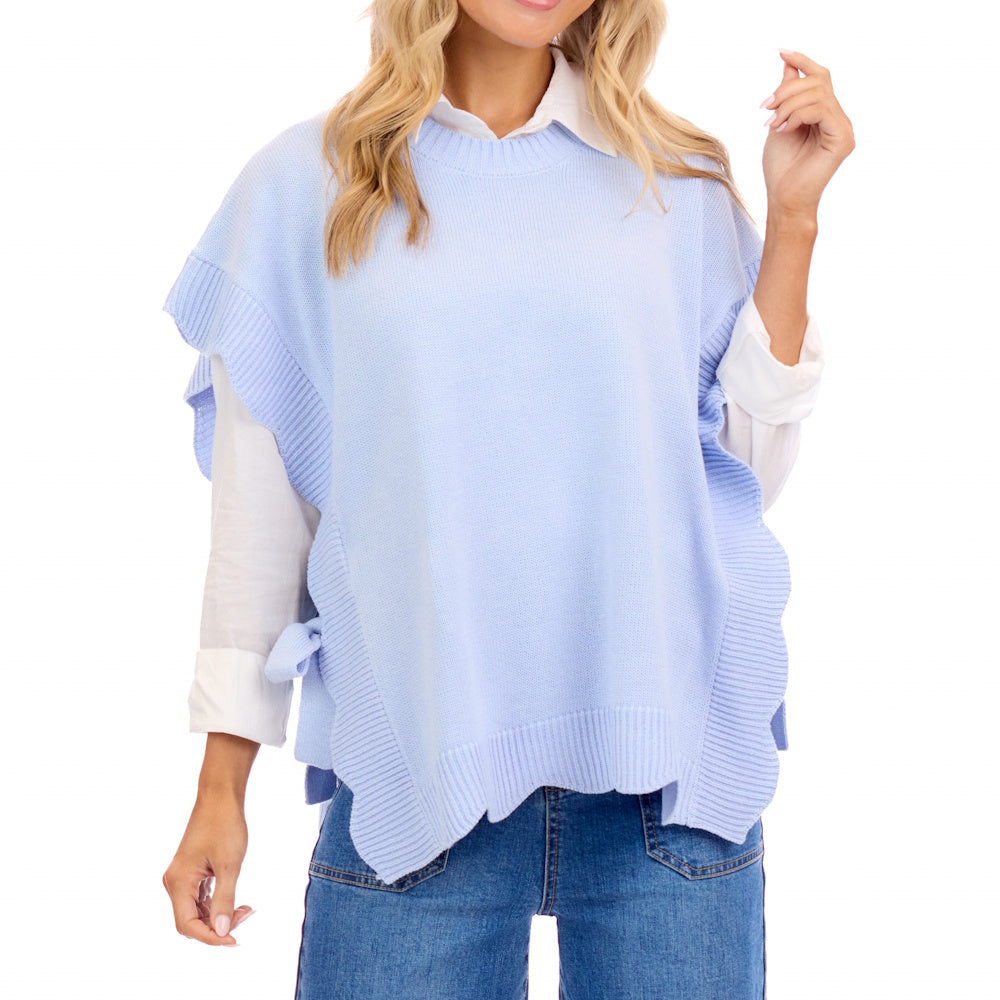 A woman wearing a light blue, short sleeve scallop-edged sweater vest over a white collared shirt and blue jeans.
