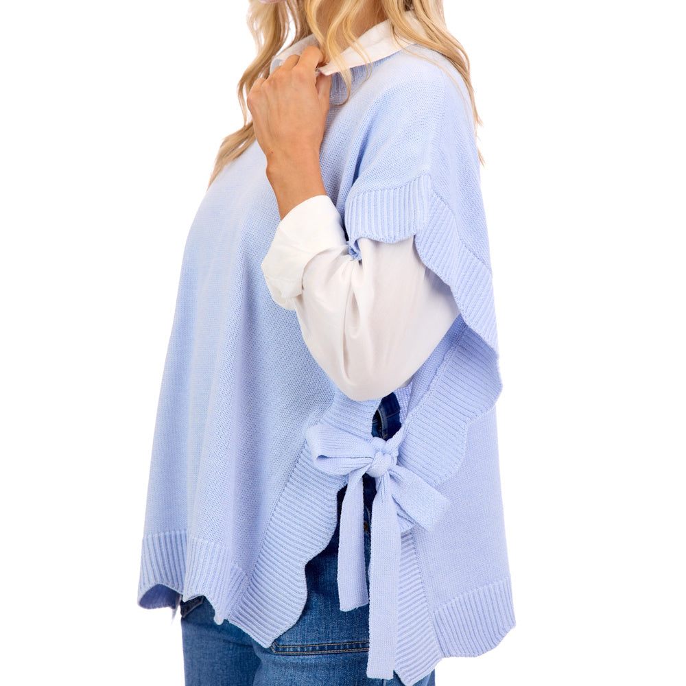 A woman wears a light blue knit sweater with scalloped edges and side bows over a white collared shirt and jeans.