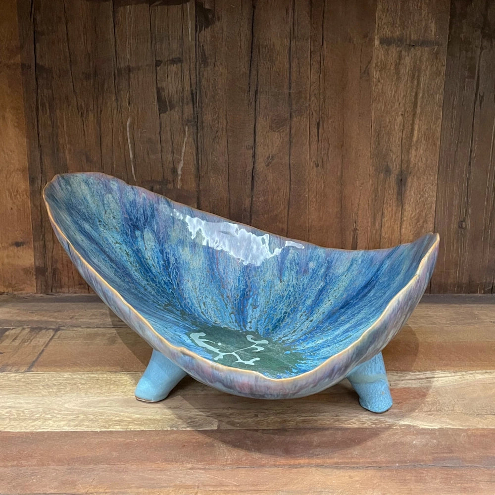 Blue stoneware footed organic shaped bowl with a reactive drip glaze in shades of teal and indigo on a wooden shelf.