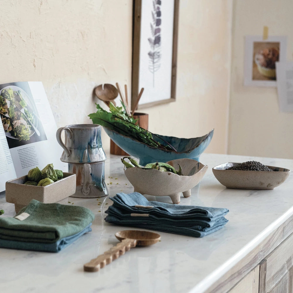 Rustic organic stoneware bowls, mugs, and linens displayed on a marble countertop with fresh greens and a cookbook.