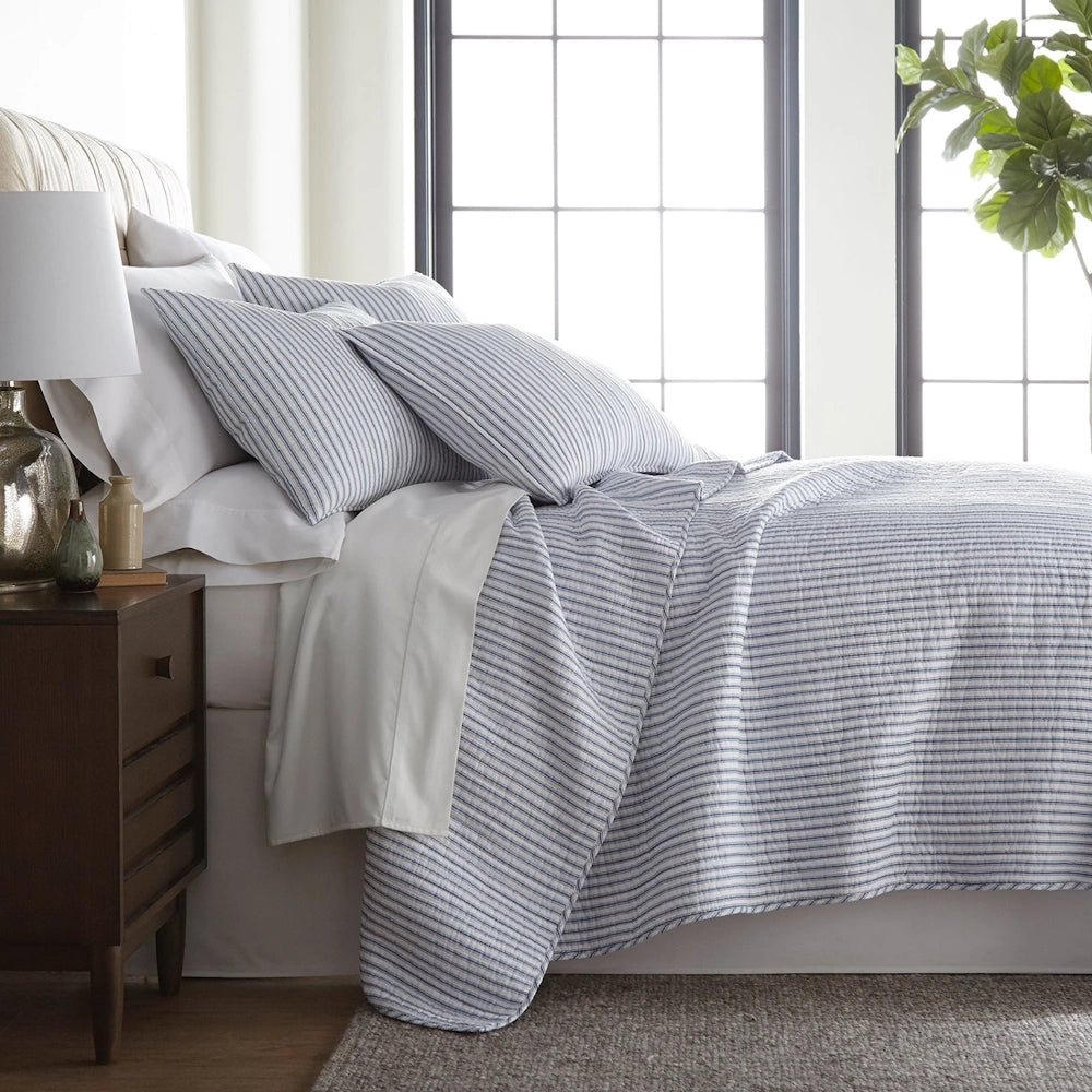 Blue and white striped quilt set on a bed with matching pillows, a wooden nightstand, and a window background.