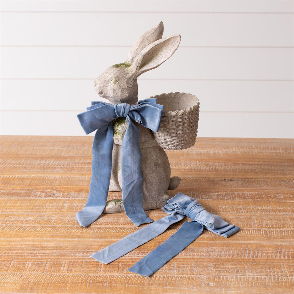 Stone rabbit figurine with blue velvet bow and basket on wooden table.