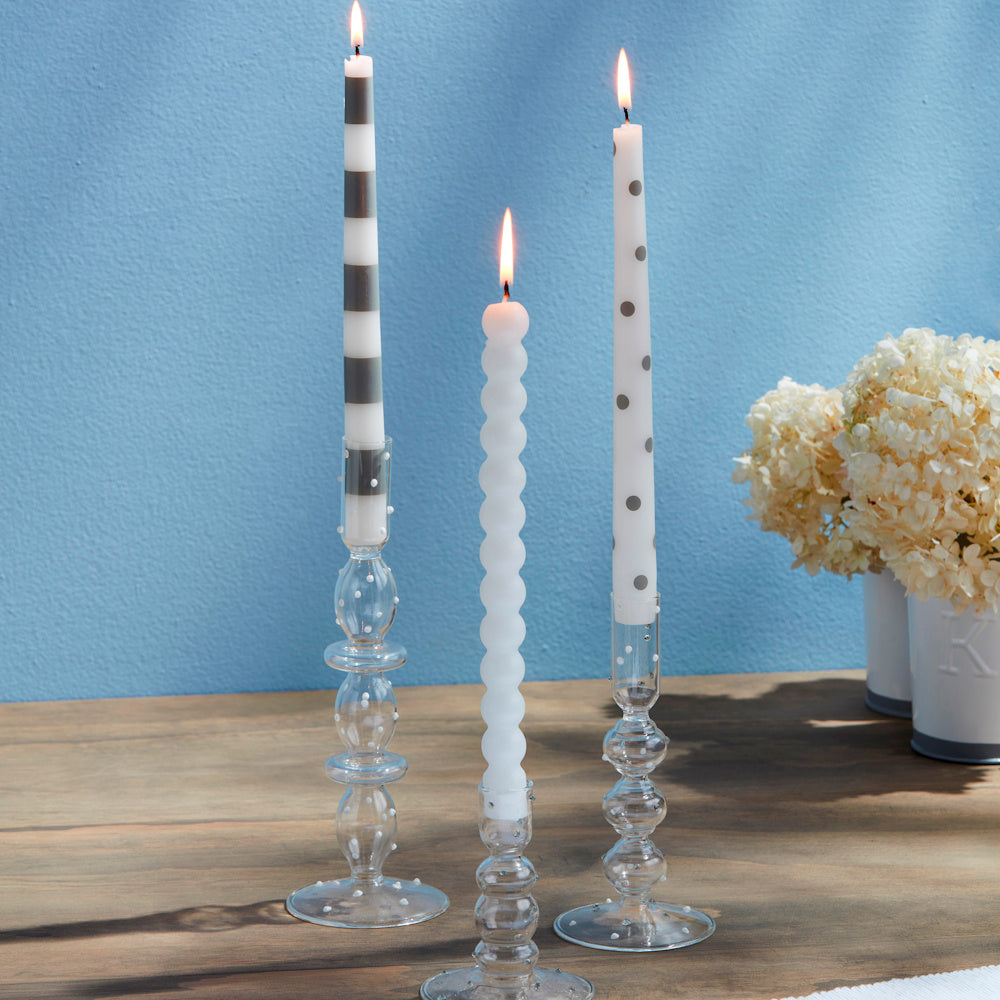 Three lit decorative taper candles in glass holders on a wooden table, featuring stripes, dots, and a bubble texture.