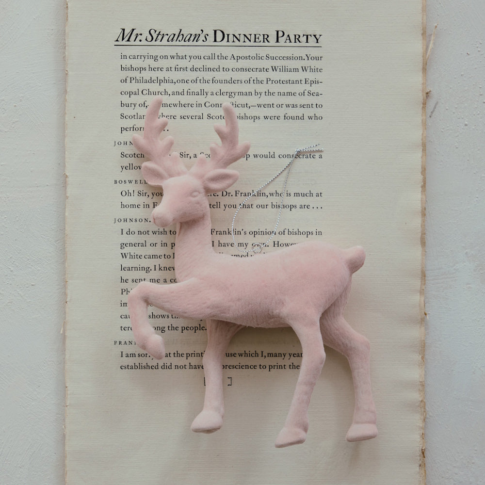 Blush pink velvet flocked deer ornament resting on a book page with a silver hanging loop.