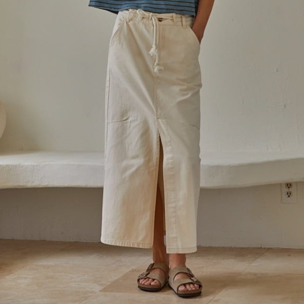 A person wearing a cream-colored denim maxi skirt with a front slit, drawstring waist, and sandals.