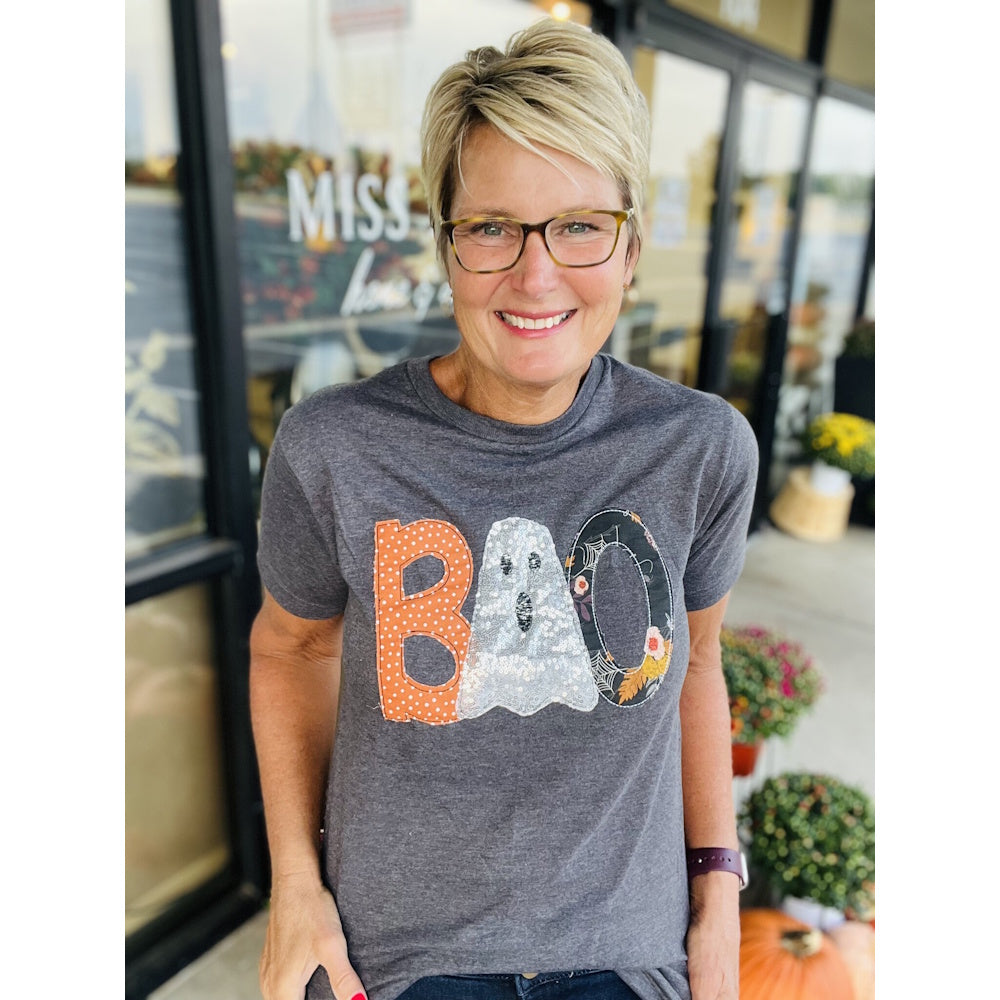 Smiling woman wearing glasses and a grey graphic t-shirt with "BOO" written in patterned letters.