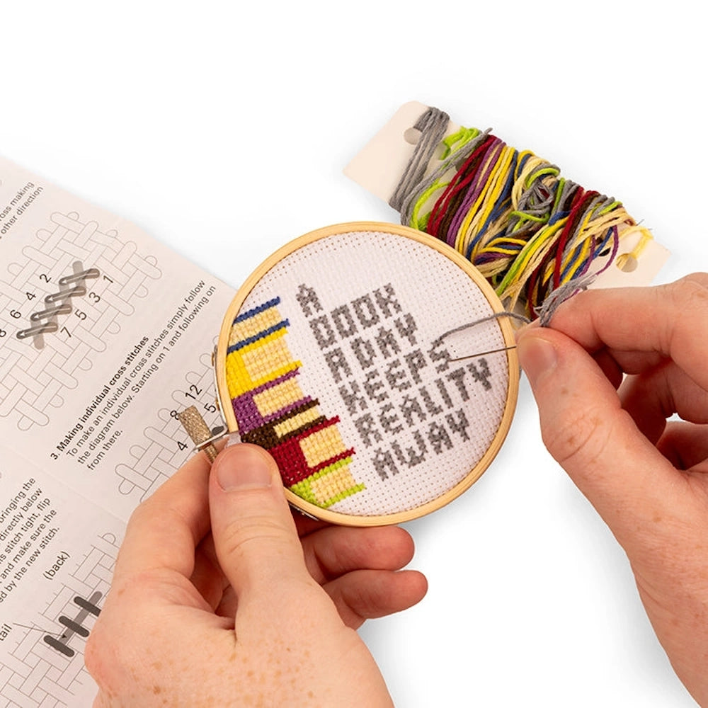 Hands stitching a mini cross-stitch of a book stack and the phrase "A book a day keeps reality away" in a wooden hoop.
