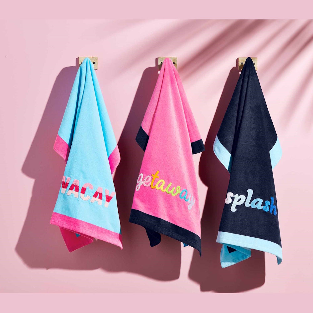 Colorful bouclé beach towels with "Vacay," "Getaway," and "Splash" embroidery hanging on a pink wall with palm shadows.