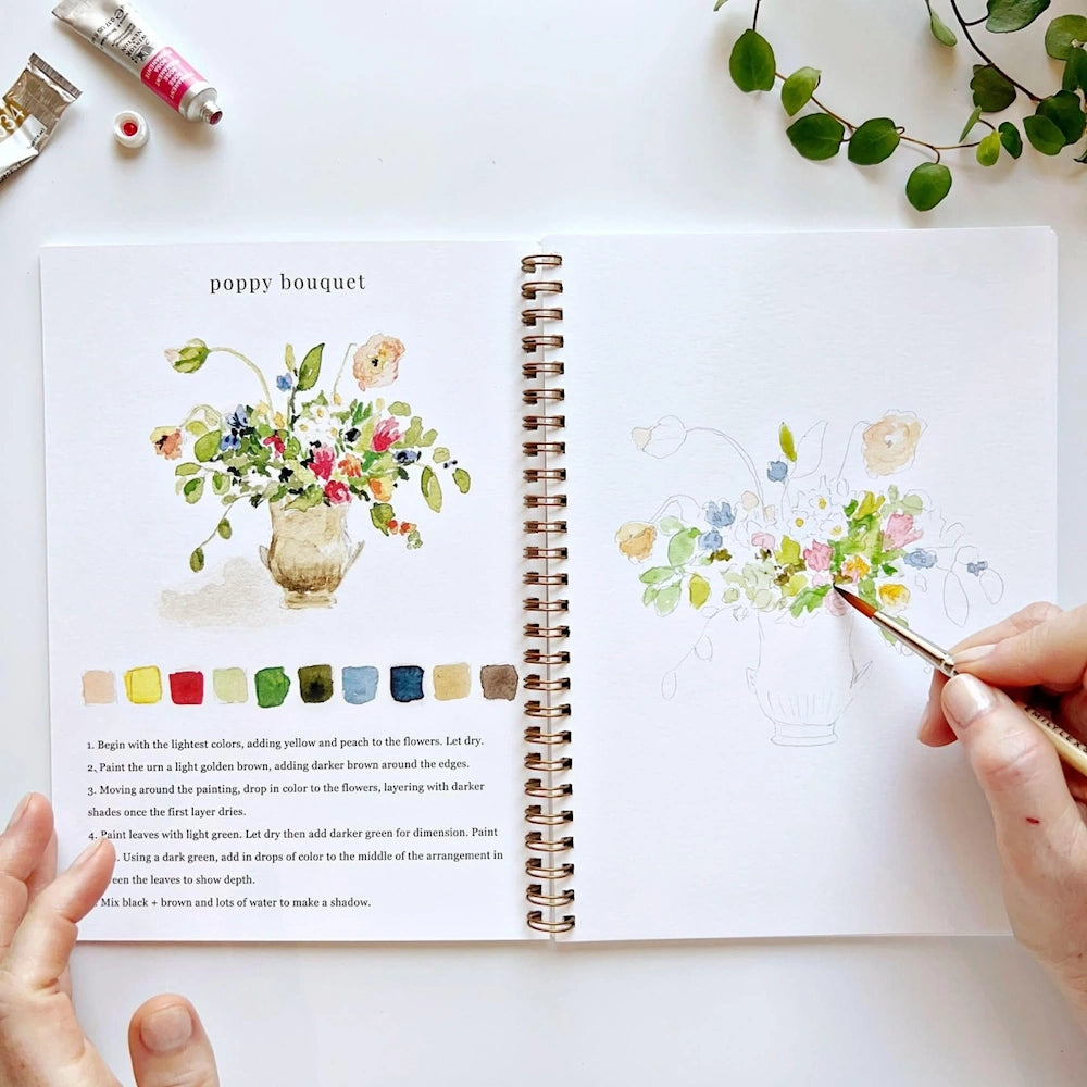 A person paints a watercolor poppy bouquet in a spiral notebook next to an instructional guide and paint tubes.